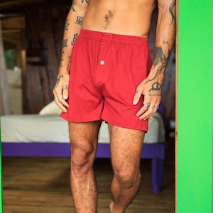 May include: Red boxer shorts with a button closure. The shorts are made of a soft fabric and are visible from the waist down. The person is standing in a doorway with a wooden floor and green door frame.