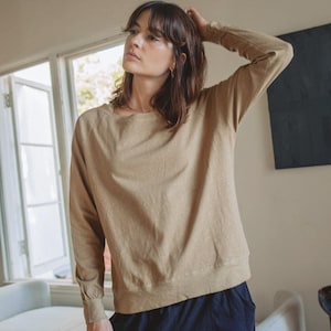 Könnte beinhalten: Ein langärmeliger beigefarbener Pullover mit Rundhalsausschnitt und gerippten Bündchen, kombiniert mit einer dunkelblauen Hose. Der Pullover hat ein schlichtes, einfarbiges Design mit einer lockeren Passform und langen Ärmeln.