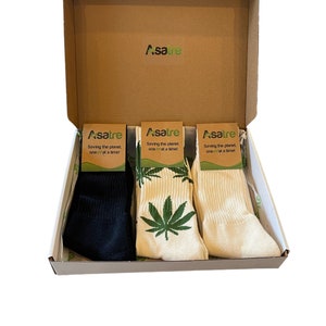 May include: A set of three pairs of socks in a cardboard box. One pair is solid black, one is cream with a green leaf pattern, and one is solid cream. Each pair has a brown paper label with the text Asatre Saving the planet, one leaf at a time.