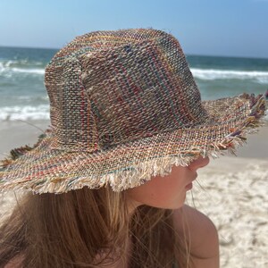 May include: A wide-brimmed straw hat with a frayed edge. The hat is made of woven straw in shades of brown, beige, and green. The hat is worn by a person with long brown hair.