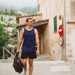 Puede incluir: Una camiseta sin mangas azul marino, pantalones cortos negros, zapatillas blancas y una bolsa de deporte marr&oacute;n. El conjunto incluye un reloj de pulsera plateado y gafas. La ropa es de estilo informal, con una camiseta sin mangas lisa, pantalones cortos deportivos y calzado blanco de corte bajo.