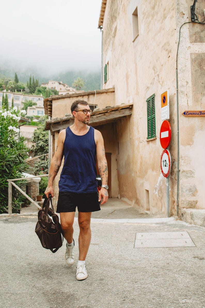 Puede incluir: Una camiseta sin mangas azul marino, pantalones cortos negros, zapatillas blancas y una bolsa de deporte marr&oacute;n. El conjunto incluye un reloj de pulsera plateado y gafas. La ropa es de estilo informal, con una camiseta sin mangas lisa, pantalones cortos deportivos y calzado blanco de corte bajo.