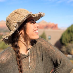 May include: A woman with long brown braids wears a light brown, wide-brimmed, woven hat with frayed edges. She is wearing a light green, long-sleeved shirt and smiling.