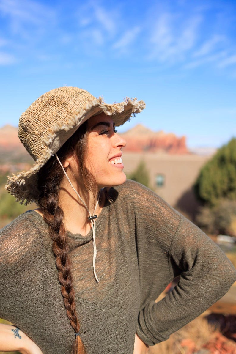 May include: A woman with long brown braids wears a light brown, wide-brimmed, woven hat with frayed edges. She is wearing a light green, long-sleeved shirt and smiling.