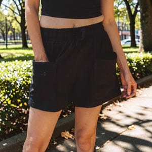 May include: Black shorts featuring an elastic waistband with a drawstring closure and two large, square patch pockets on the front. The shorts have a relaxed fit and a hem that sits above the knee, constructed from a solid black fabric.