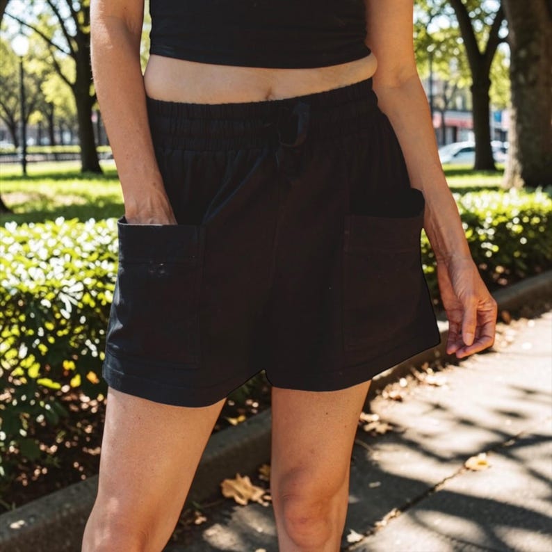 May include: Black shorts featuring an elastic waistband with a drawstring closure and two large, square patch pockets on the front. The shorts have a relaxed fit and a hem that sits above the knee, constructed from a solid black fabric.