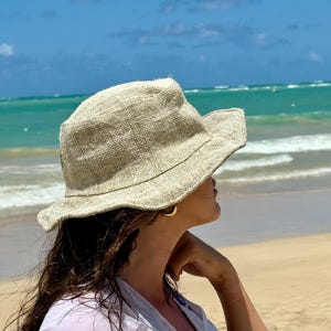 May include: A beige, wide-brimmed sun hat with a textured weave. The hat is worn by a person on a beach, with the ocean and blue sky in the background. The hat appears to be made of a natural fiber.