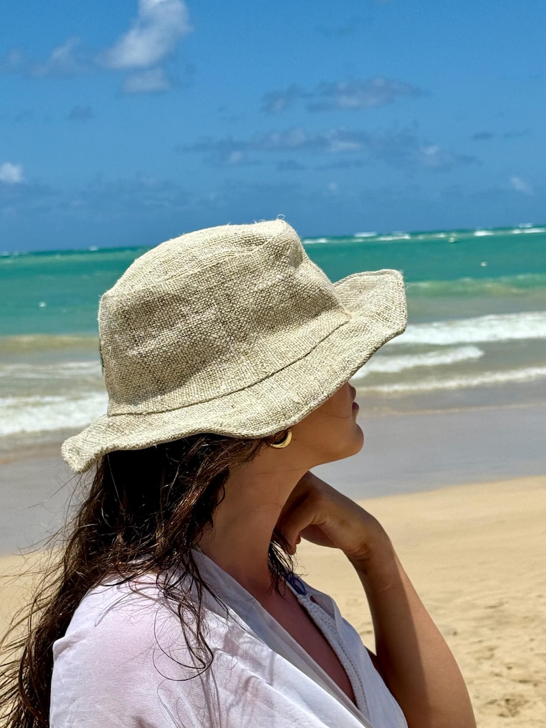 May include: A beige, wide-brimmed sun hat with a textured weave. The hat is worn by a person on a beach, with the ocean and blue sky in the background. The hat appears to be made of a natural fiber.