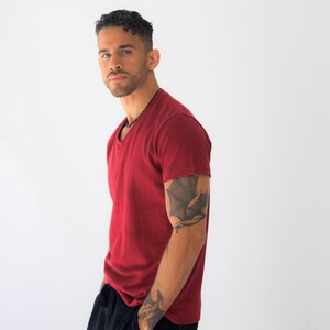 May include: A short-sleeved, deep red V-neck t-shirt with a textured fabric finish. The shirt features a standard fit with a straight hemline, providing a clean and simple look. The garment is paired with solid black pants, completing the outfit.