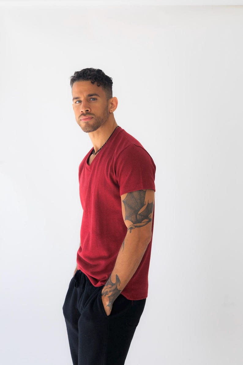 May include: A short-sleeved, deep red V-neck t-shirt with a textured fabric finish. The shirt features a standard fit with a straight hemline, providing a clean and simple look. The garment is paired with solid black pants, completing the outfit.