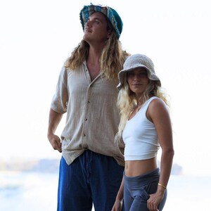 May include: A person wearing a teal and multicolored frayed bucket hat, a beige button-down shirt, and blue trousers, alongside another person wearing a beige knit bucket hat, a white tank top, and dark gray leggings.