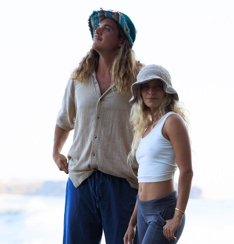 May include: A person wearing a teal and multicolored frayed bucket hat, a beige button-down shirt, and blue trousers, alongside another person wearing a beige knit bucket hat, a white tank top, and dark gray leggings.