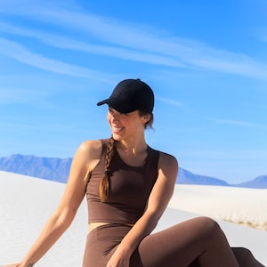 May include: A solid black baseball cap paired with a dark brown sleeveless cropped top and matching high-waisted leggings. The outfit features a simple, fitted design with a clean silhouette, worn in a bright outdoor setting with clear blue skies.
