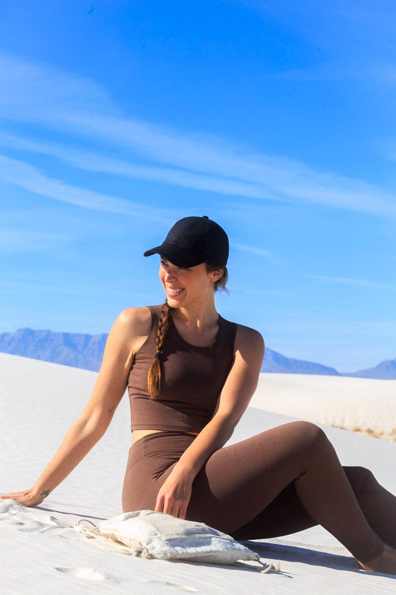May include: A solid black baseball cap paired with a dark brown sleeveless cropped top and matching high-waisted leggings. The outfit features a simple, fitted design with a clean silhouette, worn in a bright outdoor setting with clear blue skies.
