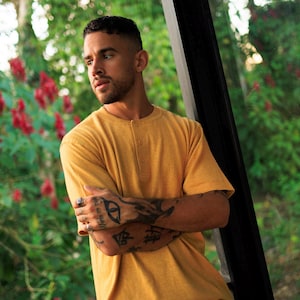 May include: Mustard yellow Henley shirt with short sleeves and three buttons. The shirt is paired with light-colored pants. The person in the image has tattoos on their arms and is leaning against a window frame, with a green background.