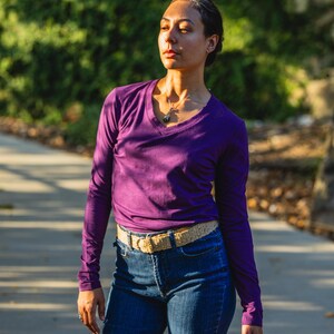 May include: A woman wearing a purple long-sleeved shirt with a v-neck and a brown belt. She is wearing blue jeans and has a necklace with a pendant.