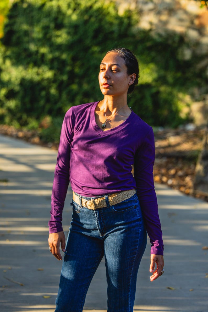 May include: A woman wearing a purple long-sleeved shirt with a v-neck and a brown belt. She is wearing blue jeans and has a necklace with a pendant.