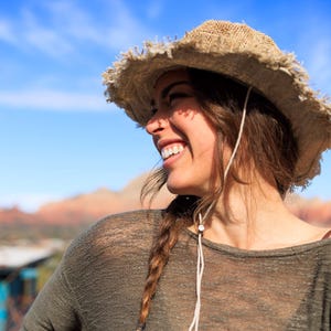 May include: A woman wearing a brown straw hat with a braided hairstyle, a green shirt, and a beaded bracelet. She is smiling and looking to the right.