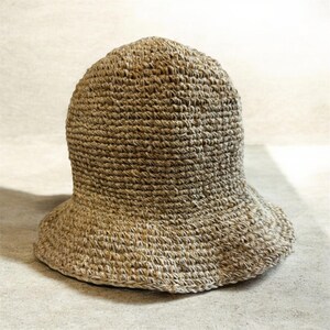 May include: A tan-colored bucket hat constructed from a thick, textured, woven fiber material. The hat features a rounded crown and a wide, downward-sloping brim, showcasing a consistent, dense crochet-like stitch pattern throughout the entire piece.