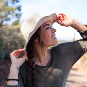 May include: A beige sun hat with a wide brim. The hat is made of a woven material and has a dark green accent. The person is wearing a long-sleeved olive green shirt. The background is a sunny outdoor scene with trees and a blue sky.