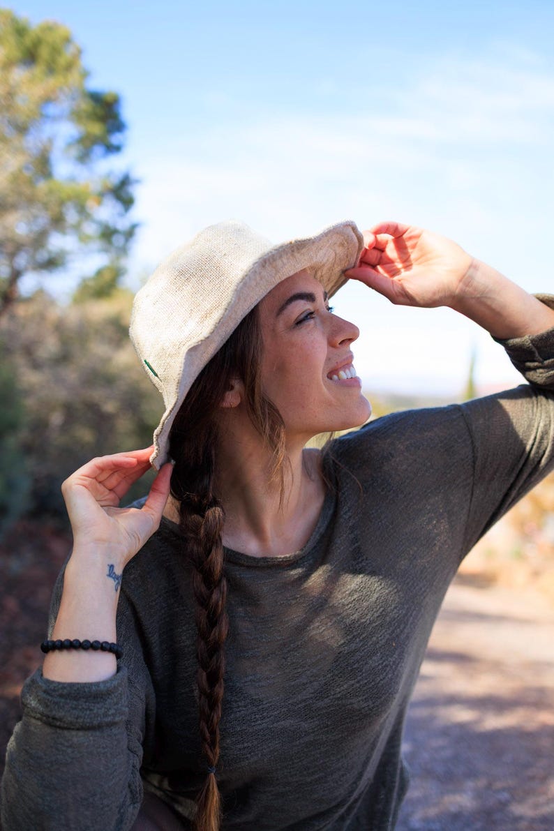 May include: A beige sun hat with a wide brim. The hat is made of a woven material and has a dark green accent. The person is wearing a long-sleeved olive green shirt. The background is a sunny outdoor scene with trees and a blue sky.