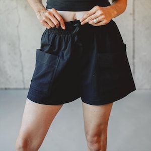 May include: Black shorts featuring an elasticized waistband with a drawstring closure and two large, square-shaped patch pockets on the front. The shorts have a relaxed fit and a straight hemline, providing a simple and functional design.