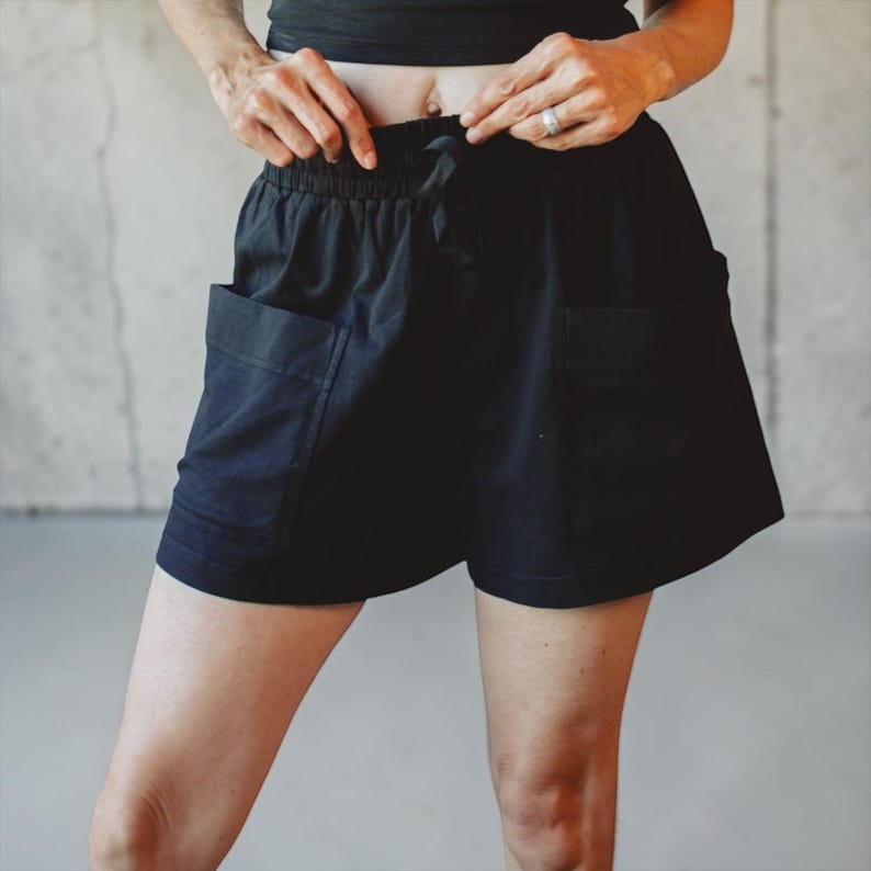 May include: Black shorts featuring an elasticized waistband with a drawstring closure and two large, square-shaped patch pockets on the front. The shorts have a relaxed fit and a straight hemline, providing a simple and functional design.
