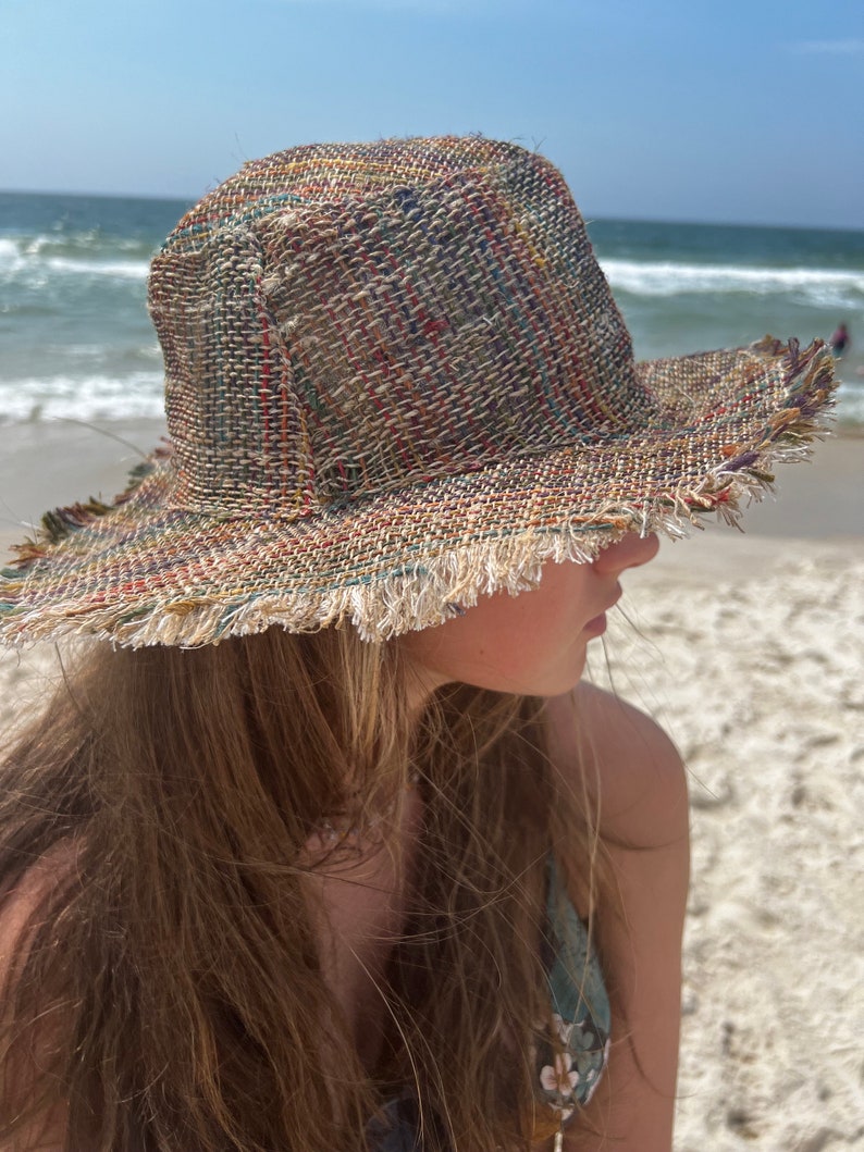 May include: A wide-brimmed straw hat with a frayed edge. The hat is made of woven straw in shades of brown, beige, and green. The hat is worn by a person with long brown hair.
