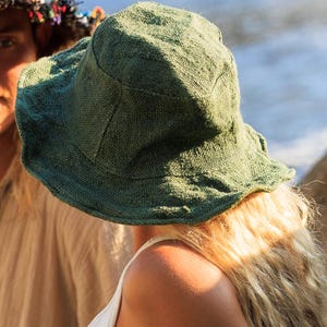 May include: A green, wide-brimmed hat made of textured fabric. The hat is worn by a person with long, blonde hair and a white tank top. The hat's brim is slightly wavy.