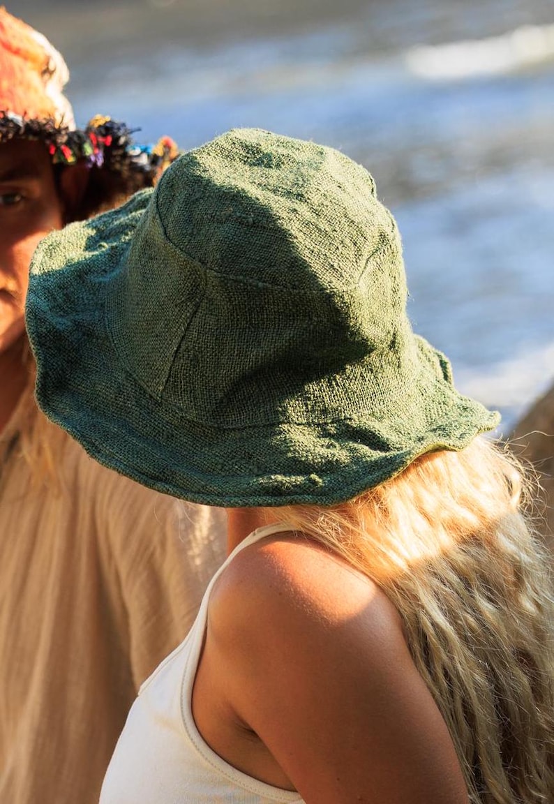 May include: A green, wide-brimmed hat made of textured fabric. The hat is worn by a person with long, blonde hair and a white tank top. The hat's brim is slightly wavy.