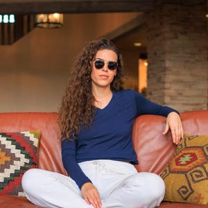 May include: A woman wearing a navy blue long-sleeved shirt and white pants sits on a brown leather couch. She is wearing sunglasses and has long curly hair.