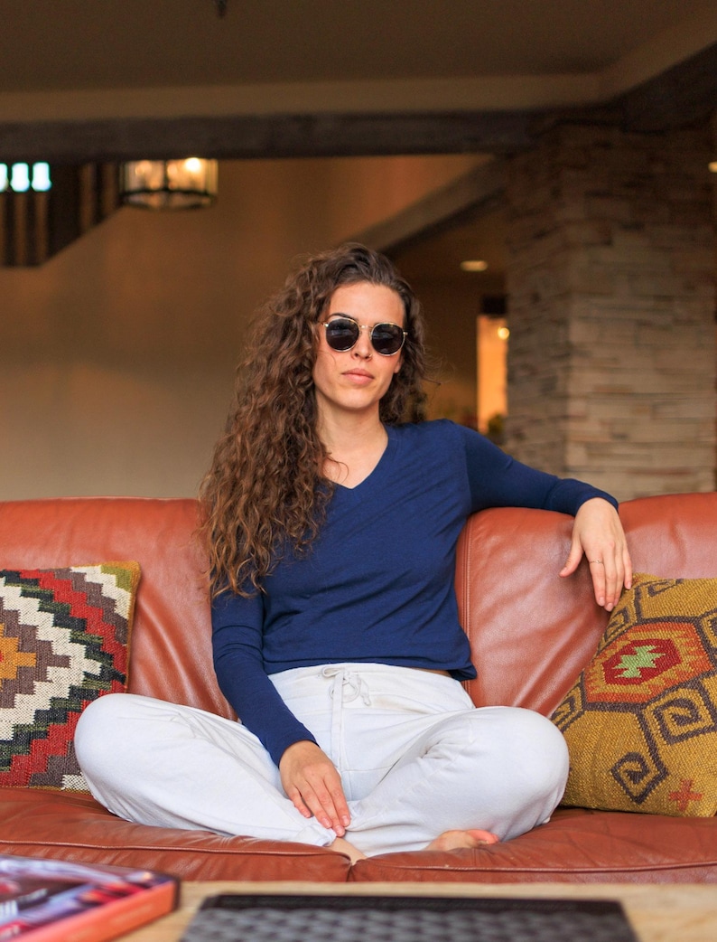 May include: A woman wearing a navy blue long-sleeved shirt and white pants sits on a brown leather couch. She is wearing sunglasses and has long curly hair.