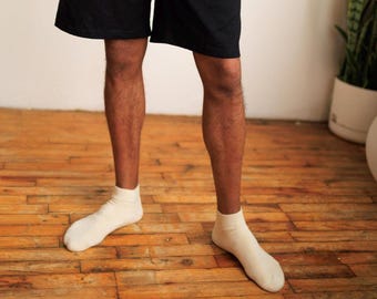 Può includere: Un paio di calzini alla caviglia color bianco sporco indossati con pantaloncini neri. I calzini presentano un bordo a coste sulla parte superiore e una struttura in maglia liscia che copre i piedi, arrivando appena sopra la caviglia.