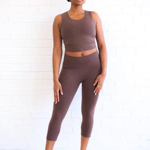 May include: A dark brown sleeveless crop top and matching dark brown cropped leggings. The set features a solid color and a fitted silhouette. The top has a round neckline, and the leggings have a high waistband that sits above the hips.