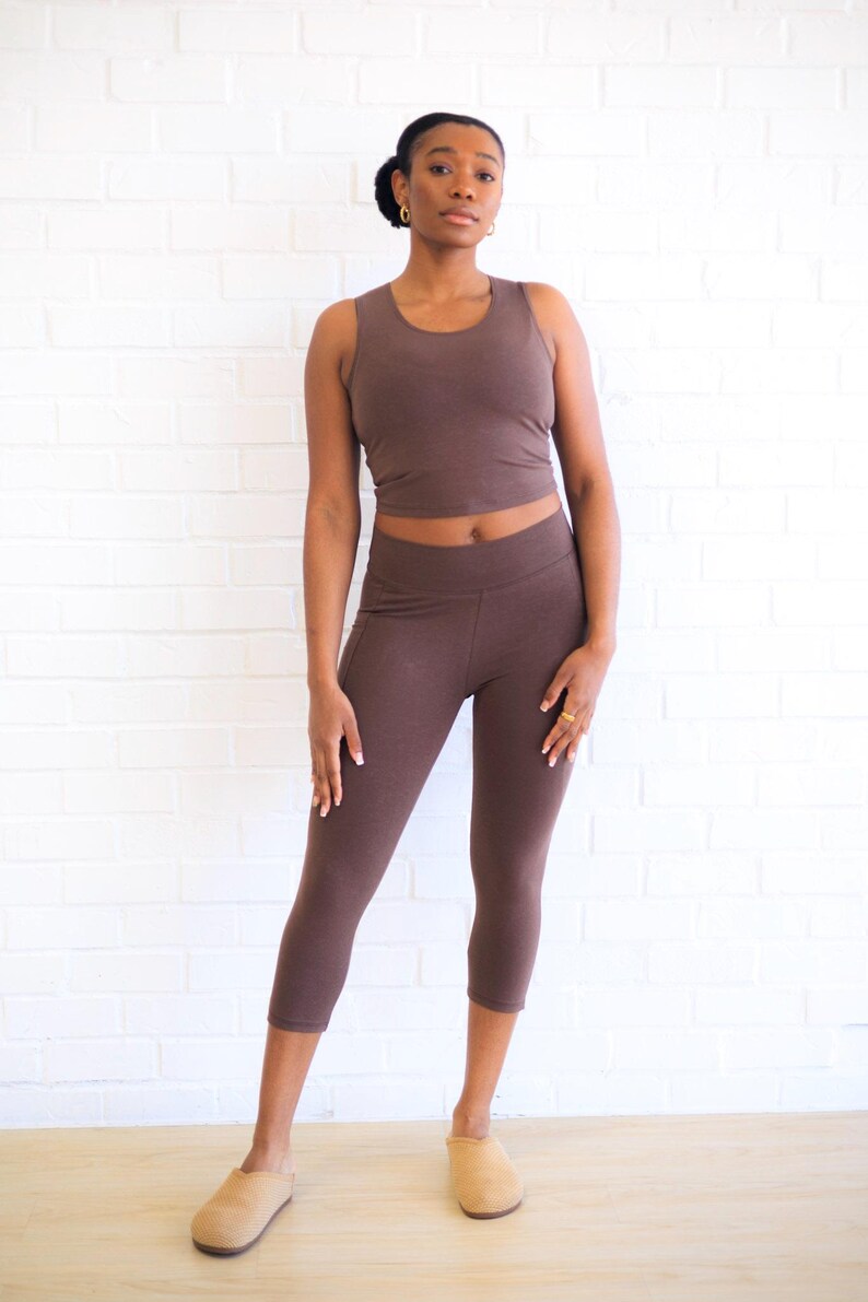 May include: A dark brown sleeveless crop top and matching dark brown cropped leggings. The set features a solid color and a fitted silhouette. The top has a round neckline, and the leggings have a high waistband that sits above the hips.
