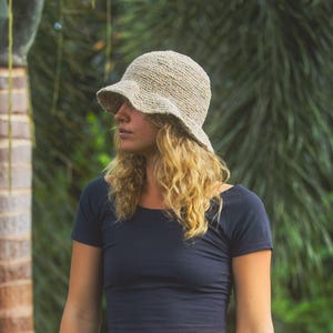 May include: A beige crochet bucket hat with a textured, woven appearance and a slightly flared brim. The hat features a natural, fibrous material construction with a visible knit pattern, providing a structured yet flexible shape for the head.