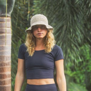 May include: A beige textured bucket hat with a wide brim and a rounded crown, paired with a dark charcoal short-sleeved crop top. The hat features a woven, open-knit construction, while the top has a simple scoop neckline and a fitted silhouette.