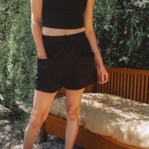 May include: A black sleeveless crop top with a scoop neckline paired with black shorts featuring an elastic waistband, a drawstring, and two large front patch pockets. The shorts have a relaxed fit and reach mid-thigh, providing a simple look.