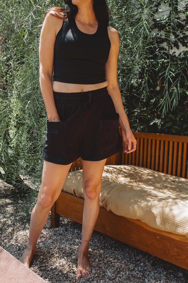 May include: A black sleeveless crop top with a scoop neckline paired with black shorts featuring an elastic waistband, a drawstring, and two large front patch pockets. The shorts have a relaxed fit and reach mid-thigh, providing a simple look.
