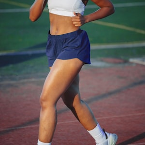 Könnte beinhalten: Ein weißer Racerback-Sport-BH, dunkelblaue Sportshorts, weiße Crew-Socken und weiße Sportschuhe. Das Ensemble besticht durch ein klares, funktionales Design, wobei die Shorts bis zur Mitte des Oberschenkels reichen und die Schuhe eine sichtbare Luftdämpfung im Fersenbereich aufweisen.