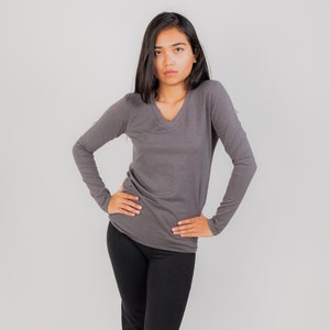 May include: A woman wearing a gray long-sleeved v-neck shirt and black pants. She is standing with her hands on her hips.