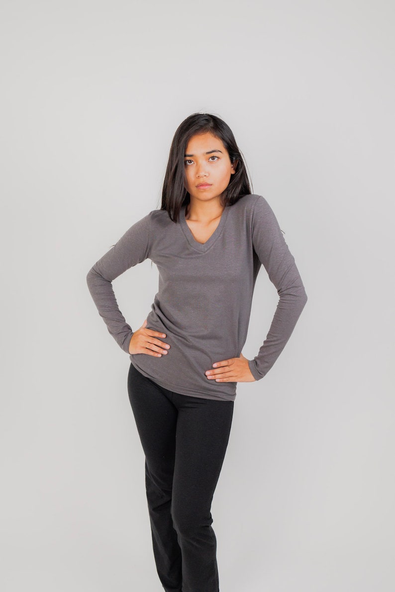 May include: A woman wearing a gray long-sleeved v-neck shirt and black pants. She is standing with her hands on her hips.