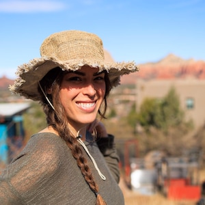 May include: A woman with long brown hair braided on one side is wearing a brown, wide-brimmed, woven hat with frayed edges. She is smiling and looking at the camera. She is wearing a green, long-sleeved shirt.