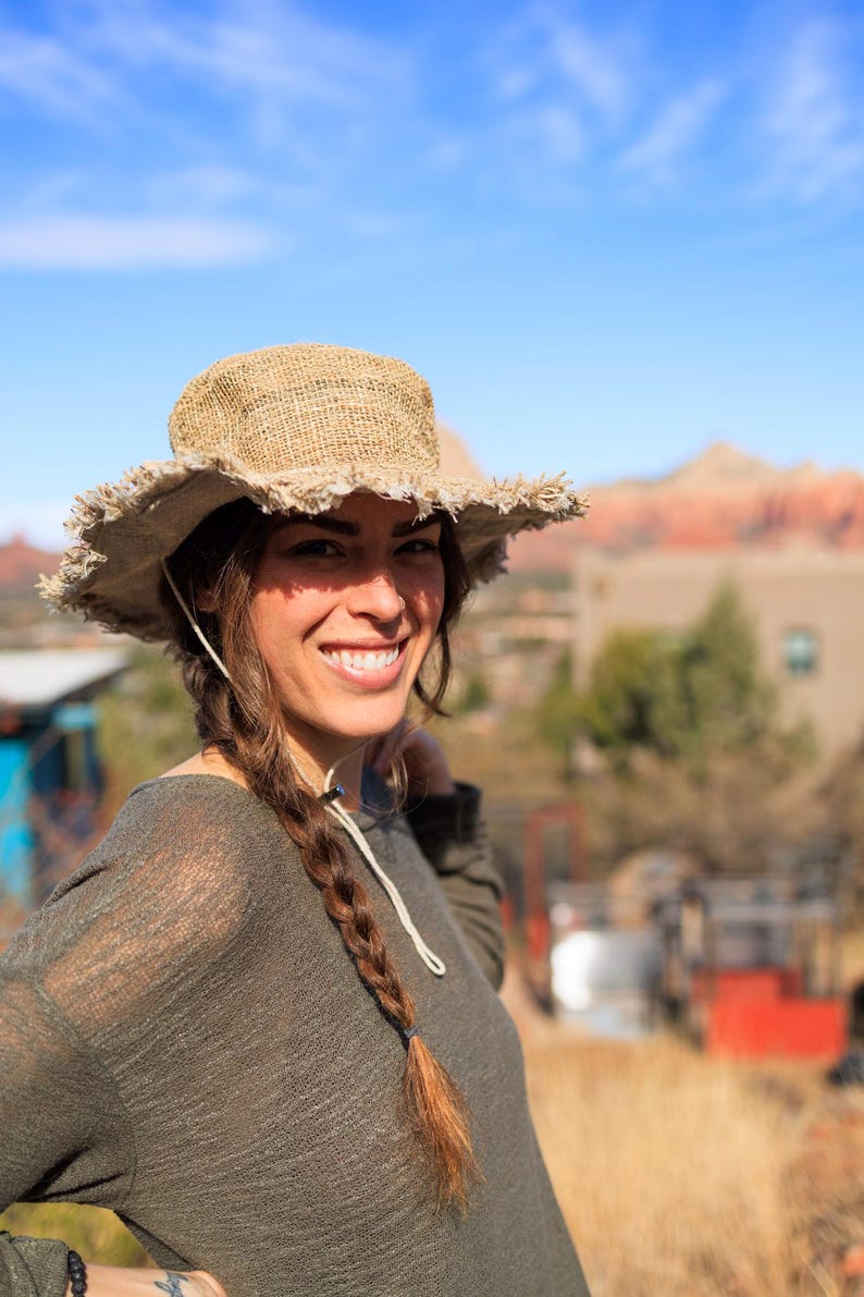May include: A woman with long brown hair braided on one side is wearing a brown, wide-brimmed, woven hat with frayed edges. She is smiling and looking at the camera. She is wearing a green, long-sleeved shirt.