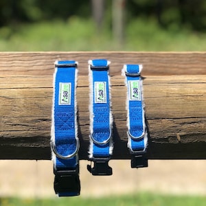 May include: Three blue dog collars with white faux fur trim and black plastic buckles. Each collar has a silver metal D-ring and a rectangular label with the text "Asare" in white and green. The collars are displayed on a wooden surface.