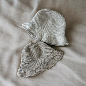 May include: Two crochet bucket hats in neutral tones. The top hat is a light cream color, while the bottom hat is a slightly darker beige. Both hats feature a rounded crown and a short, flexible brim with a consistent, dense stitch pattern.
