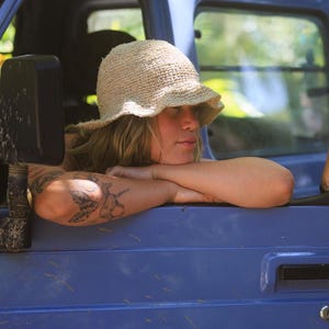 May include: A beige, textured, woven bucket hat with a soft, flexible brim. The hat features a simple, open-knit construction with a rounded crown, providing a natural, neutral-toned appearance that sits comfortably on the head.