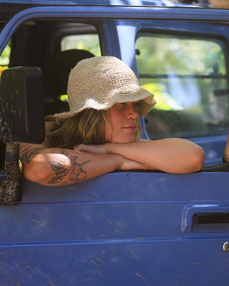 May include: A beige, textured, woven bucket hat with a soft, flexible brim. The hat features a simple, open-knit construction with a rounded crown, providing a natural, neutral-toned appearance that sits comfortably on the head.