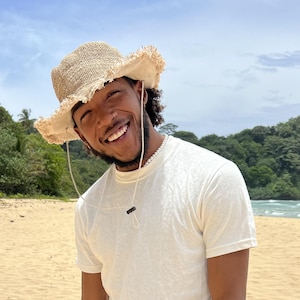 May include: A person wearing a white t-shirt and a light brown straw hat smiles at the camera on a beach. The person is standing in front of a sandy beach with green trees and a blue ocean in the background.