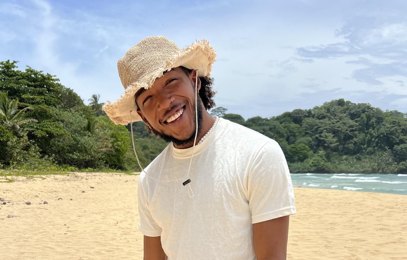 May include: A person wearing a white t-shirt and a light brown straw hat smiles at the camera on a beach. The person is standing in front of a sandy beach with green trees and a blue ocean in the background.